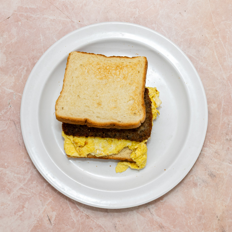 Breakfast sandwich with toasted bread, sausage patty, and scrambled eggs on a white plate.
